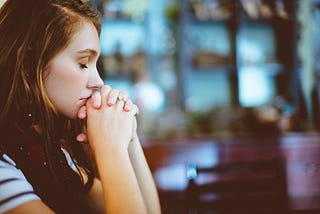 Lady sits with her eyes closed and hands clasped like she is in prayer.