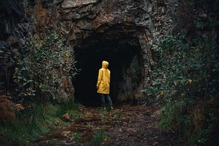 A woman in a strong yellow raincoat standing at the entrance of a large cave looking at its darkness. We all go though dark moments. But can come out strengthened.