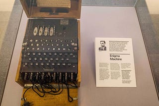 Museum display shows Enigma coding machine, black with keys like a typewriter, on left and info card on right.