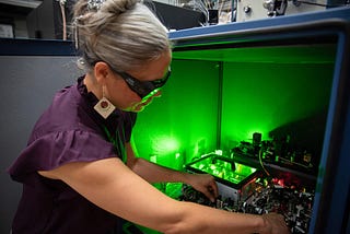 A researcher wearing safety glasses reaches into a box of circuitry and other equipment, which emits a green glow.