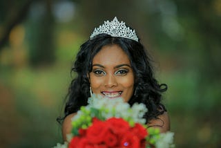 A smiling, beautiful young woman with long dark hair, wearing a tiara, and holding up red and white roses, as if at a prom.