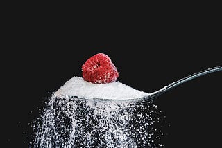 Close-up of an over-flowing silver teaspoon of sugar with a red raspberry on top, against a black background.
