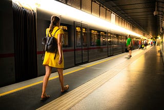 A train on the platform. A passenger in a yellow dress walks along the platform. In the end, more people are waiting.