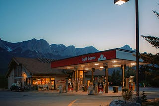 Self-serve gas station bearing a Canadian maple leaf motif, with a shop/café standing behind the forecourt. Beyond them is a mountain range. There is a clear sky but the sun has just gone down and both the station’s forecourt and the shop have their lights on.