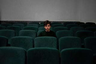 An adolescent white boy with swoopy brown hair wearing a black long sleeve shirt sits alone in a room of velvet green theatre chairs with an angry expression.