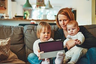 A mother sits on the couch holding her baby. Her toddler sits next to her. They are watching a tablet. Mother is pondering her next Medium article.