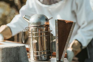 Photo of a beekeeper in white overalls and veil in the background, and a close-up of a smoker in the foreground.