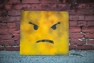 Large yellow square frowny face leaning up against a brick wall.