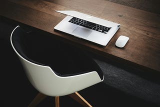 An extremely neat workplace — white chair at a dark-wood desk. A white laptop and a white mouse neatly laid out on the desk.