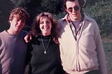 Three siblings standing side by side looking at the camera with trees in the background. Two brothers on either side of their sister in the middle. It’s 1981. The brothers are 19 and 21. The sister is 17.