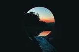 a black tunnel opens onto sunset and trees over water