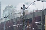 An artsy-crafty photo — almost in black and white, but with sepia highlights, of a dreary, soggy, foggy, Seattle day — antique lamp posts in the foreground, then viaduct overpass, with misty high-rise apartments in the background.