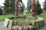 A modest marble cross engraved SEALTH marks the final resting place of Chief Seattle. The cross is butressed by two large cedar columns carved in tribal motifs and painted red and black. The grave site is enclosed by a low cement wall engraved in English and the native dialect with excerpts from a speach attributed to Chief Serattle.