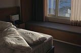 A bed with one white pillow, white bedspread, and white sheets. Bed is near a window with tan curtains. There is a lamp with a square white lampshade.