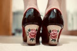 The back of the flamenco shoes of a woman, whose white ankles you can see. The heels are black leather and the heels wooden with red roses and spiky leaves peeping out the sides.