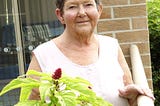 Elderly woman looking at the camera with a gentle smile on her face. One hand rests on a balcony railing and a plant with a red flower is in the forefront.