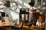 A woman walking in her kitchen with a cup of coffee in her hand, with her dog beside her.