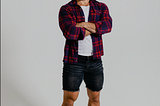 Youtuber Alex Homorzi in an unbuttoned red plaid shirt, white t-shirt underneath, black denim shorts, white socks and crocs