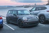A Brown Mini Cooper Clubman with racing stripes and a sunset in the backround