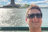 Man wearing sunglasses on boat with Statue of Liberty in background.