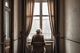 Lonely elderly woman soft brown wool sweater is sitting on a chair in front of the window facing us with her hutched back. cloudy day outside. You see gre colors. All walls are in vintage slightly ripped wallpaper pink-greay colors.