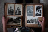 Sepia-toned aged photo journal held by pink hands. Open to black and white photograph, memories removed by a generation or two. What happened between two people so long ago? What happens to an unconfirmed affair?