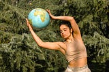 color photograph of a woman standing outside in front of pine trees with her body beautifully arched to the side while holding a globe