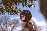 color photograph of a woman with brown hair and glasses holding a mirror and gazing calmly at her reflection while outside on a clear day