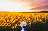 A woman in a white dress raises her arms in a field of sunflowers at sunset