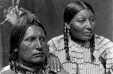 A Native American husband and wife, Mr. and Mrs. American Horse, look at something out of frame. She lays her hand on his shoulder.