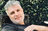 Headshot of a relaxed man with gray hair wearing a black t-shirt sitting outside in a garden with a green bush behind him.