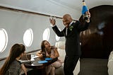 A well-dressed man dancing on a private jet while holding a bottle of champagne and two champagne flutes, with two young women sitting and smiling at him.