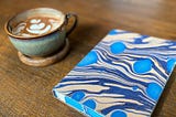 A marbled blue journal next to a cup of coffee with flower latte art on a wooden table