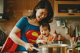 A Chinese woman wearing a superhero costume and laughing together with her baby as she prepares food in a kitchen