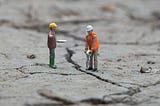 Two construction worker action figures with hardhats jackhammering into the ground