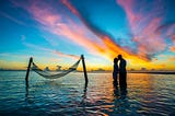 A couple in silhouette kissing while standing in thigh-deep water with a spectacular sunset in the background.