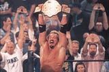 A blond man, Diamond Dallas Page, holds a golden belt over his head. Behind him, a crowd cheers him on.