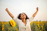 A woman in a sunflower field smiles and holds her arms skyward with positivity.