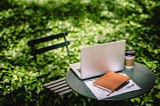 small table with black chair, laptop, journal, and coffee cup on it.