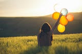 Back of a long-haired woman holding up balloons in meadow at the edge of hills