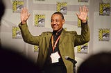 Giancarlo Esposito waves to a crowd with both hands. He’s wearing a green jacket and standing in front of a Comic-Con backdrop.