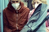 man and woman wearing medical masks sitting on a train with a child in a carseat in front of them