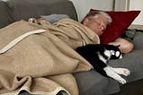 Man asleep on sofa with black and white cat sleeping next to him.