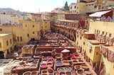 View of open-air tannery with vats of different colored dyes in the middle of the photo and yellow buildings on each side. Brown hides are drying on the walls of the building on the right side.