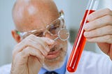 Photo of an older man in a lab coat examining a test tube filled with red liquid using a magnifying glass. That is to say, there is no actual science taking place in the photo, just the veneer of “science-ness.”