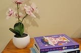 A stack of books by Lia Louis placed on a wooden table beside a pot of orchids