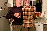 Two men stand in a kitchen with their arms around each other, holding up glasses of beer.