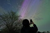 Silhouette of a person taking a phote of the sky with their phone. The sky is ablue with stripes of green and purple