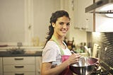 Woman standing in a kitchen while wearing pink apron and holding mixing bowl and whisk, smiling at camera