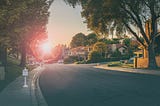 A view looking down a tree-lined road in a suburban neighborhood with houses on either side and the sun just above the horizon